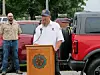 Man speaking at a wooden podium outdoors with two people and parked vehicles behind him.
