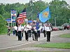 A small group in uniform marches down a road carrying American and blue organizational flags.