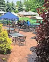 Outdoor café patio with metal tables and chairs, umbrellas, and a few seated patrons amid landscaped plants.
