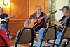 Three men seated indoors play acoustic guitars in a room with wood-paneled walls and empty chairs in the foreground.