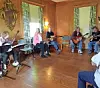Several seated musicians perform in a wood-paneled room with guitars and sheet music stands.