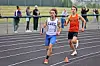 Two runners compete on a track, with spectators visible behind them.