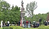 People gathered around a tall monument in a park with trees and a speaker on a stand nearby.
