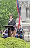 Person speaking at a podium outdoors during a memorial ceremony beside seated uniformed service members and flags.