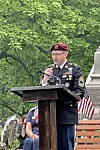 Man in military uniform speaking at a podium outdoors with trees and American flags behind him.