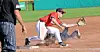 Baseball player in red fields a grounder near first base while a runner slides in.