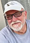 Close-up of a man wearing glasses and a white logo cap against a wood-paneled wall.