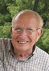 Smiling older man wearing glasses and a plaid shirt in front of green trees.