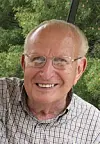 Smiling older man wearing glasses and a plaid shirt in front of green trees.