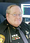 Police officer in uniform seated beside a computer monitor in an office.