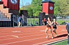 Two runners compete on a red outdoor track near stadium seating and red towers.