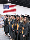 Graduates in black caps and gowns stand in line indoors beneath a large American flag.
