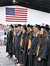 Graduates in black caps and gowns stand in line indoors beneath a large American flag.