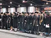 A line of graduates in caps and gowns standing indoors during a commencement ceremony.