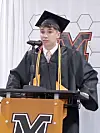 A graduate in a cap and gown speaks at a podium with a microphone.