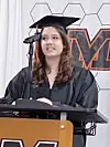 Graduate in cap and gown speaking at a podium with a microphone during commencement.