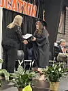 A graduate in a cap and gown shakes hands with a woman presenting a diploma at a commencement ceremony.