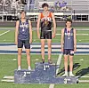 Three athletes wearing medals stand on first-, second- and third-place podium blocks at a track stadium.