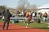 Runner in orange and black uniform on an outdoor track with people and tents in the background.