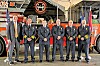 A group of firefighters and officials stand in front of a red fire truck inside a station.
