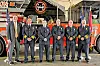 A group of firefighters and officials stand in front of a red fire truck inside a station.