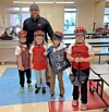 Four children in bike helmets stand with an adult inside a cafeteria-like room.