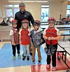 Four children in bike helmets stand with an adult inside a cafeteria-like room.