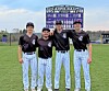 Four baseball players stand side by side on a baseball field in front of a scoreboard.