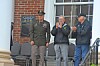 An older man claps on a porch next to a uniformed officer and another man outside a brick building.