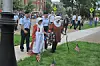 People gathered beside a wreath and small American flags near a sidewalk and grass.