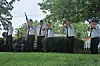 Men in white shirts and dark pants stand outdoors holding rifles with trees and bushes behind them.