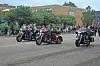 A group of motorcyclists rides in formation past people standing on the sidewalk.