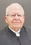 Older man with glasses wearing a dark jacket against a gray background.
