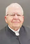 Older man with glasses wearing a dark jacket against a gray background.