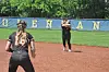 Two softball players stand on a dirt infield with an outfield wall and trees in the background.