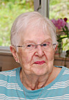 Elderly woman indoors near a window, with flowers and soft daylight behind her.