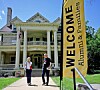 Two people stand outside a historic mansion near a yellow Welcome banner for Alumni & Families.