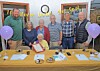 A group of adults gathered behind a table with papers, glasses, and a framed certificate in a decorated room.
