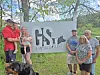 A small group stands beside a GS Swine sign outdoors near trees and grass.