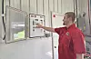 Man in a red shirt points to a wall-mounted control panel in an industrial room.
