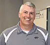 Smiling man in a gray polo shirt standing indoors with office equipment behind him.