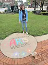 Person standing beside a chalk mural that says “medium county Arts Week” in a park.