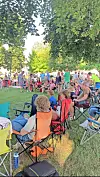 Crowd seated in folding chairs on grass under trees at an outdoor gathering.