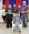 Five people pose indoors before red and blue curtains, with one seated in a white chair in front.