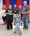 Five people pose indoors before red and blue curtains, with one seated in a white chair in front.