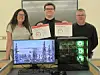 Three people hold certificates behind a desk with a monitor and a desktop computer tower.