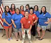 Eight women pose together indoors in red and blue T-shirts in front of a corrugated metal wall.