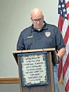 A man stands at a podium with a pledge sign, speaking beside an American flag.