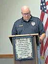 A man stands at a podium with a pledge sign, speaking beside an American flag.