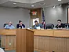 Several officials sit behind a wooden dais in a meeting room with an American flag.